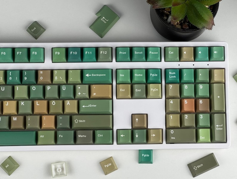 May include: A full-sized mechanical keyboard with a white frame and green, tan, and brown keycaps. The keycaps are arranged in a standard QWERTY layout, with function keys and a number pad. Several loose keycaps are scattered around the keyboard.