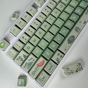 Puede incluir: Un teclado mecánico blanco con teclas personalizadas en tonos verdes y blancos. Las teclas presentan varias ilustraciones, incluyendo una rodaja de sandía, setas y plantas. El teclado tiene una distribución QWERTY estándar.