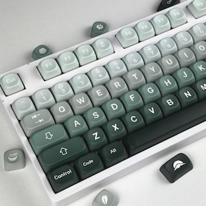May include: A white mechanical keyboard with green gradient keycaps. The keycaps transition from light green to dark green, with white lettering. Several extra keycaps with unique symbols are scattered around the keyboard.