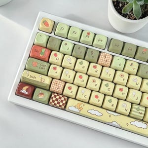 May include: A white mechanical keyboard with custom keycaps. The keycaps are a mix of light green and cream colors, with various cute illustrations of fruits, animals, and patterns. The space bar has a yellow design with clouds and a moon.