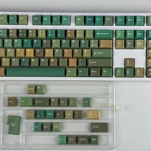 May include: A white mechanical keyboard with green and tan keycaps. The keyboard has a full layout with function keys and a number pad. A clear plastic case holds extra keycaps. The keycaps are various shades of green and tan.