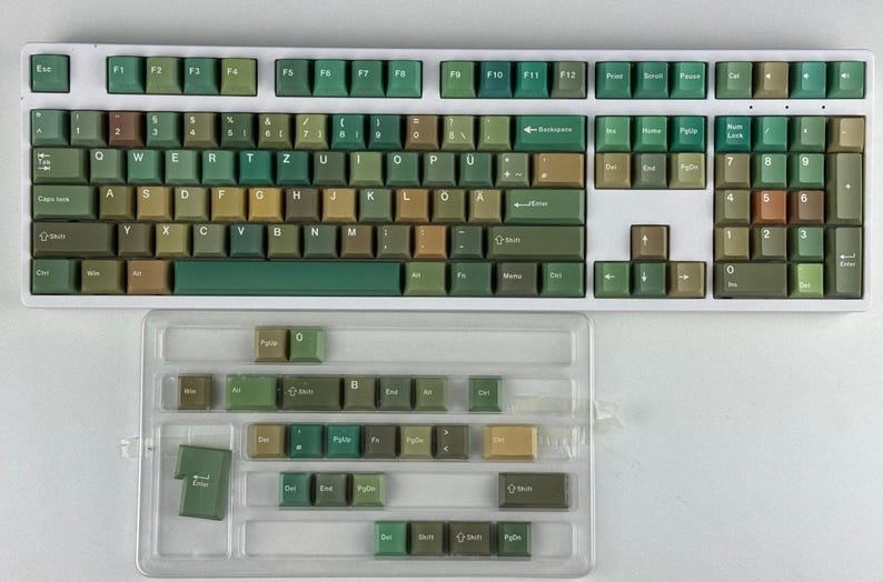 May include: A white mechanical keyboard with green and tan keycaps. The keyboard has a full layout with function keys and a number pad. A clear plastic case holds extra keycaps. The keycaps are various shades of green and tan.