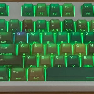 May include: A close-up of a white mechanical keyboard with green backlighting. The keycaps are a mix of colors, including green, brown, and yellow. The keyboard has function keys and number keys.
