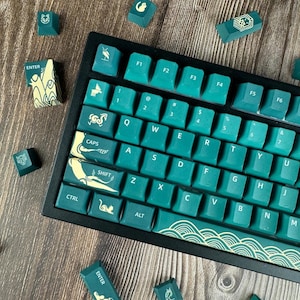 May include: A teal and black mechanical keyboard with custom keycaps. The keycaps feature white and gold designs, including waves and symbols. Several loose keycaps are scattered around the keyboard on a wooden surface.