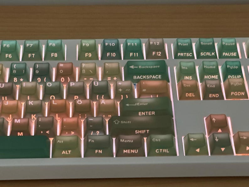 May include: A close-up of a mechanical keyboard with a white frame and multi-colored keycaps. The keycaps have a gradient color scheme, transitioning from green to brown. The keyboard features function keys, a backspace key, and other standard keys.