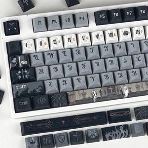 Könnte beinhalten: Eine individuelle mechanische Tastatur mit grauen und schwarzen Tastenkappen. Die Tastenkappen weisen einzigartige Symbole, römische Ziffern und stilisierte Grafiken auf. Die Tastatur hat einen weißen Rahmen und enthält zusätzliche Tastenkappen mit verschiedenen Designs.