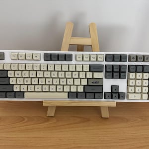 May include: A white mechanical keyboard with gray and off-white keycaps. The keyboard is displayed on a small wooden easel. The keys are arranged in a standard QWERTY layout, with function keys and a number pad.