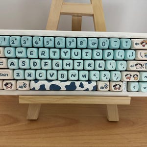 Könnte beinhalten: Eine weiße mechanische Tastatur mit hellblauen Tastenkappen. Die Tasten sind mit weißen Buchstaben und Zahlen beschriftet. Einige Tasten zeigen Illustrationen zum Thema Kuh. Die Tastatur wird auf einer kleinen Holzstaffelei präsentiert.