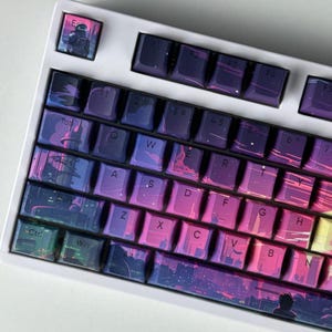 May include: A white mechanical keyboard with custom keycaps. The keycaps feature a cityscape design in shades of purple, pink, and blue. The "Escape" key has an astronaut graphic. The keyboard has standard QWERTY layout.