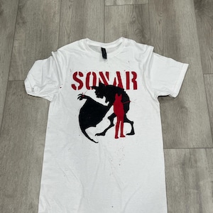 Könnte beinhalten: Weißes T-Shirt mit dem Wort "SONAR" in roten Großbuchstaben über einer schwarzen und roten Grafik. Die Grafik zeigt ein fledermausartiges Wesen und eine rote Figur. Das T-Shirt ist aus weichem Material.