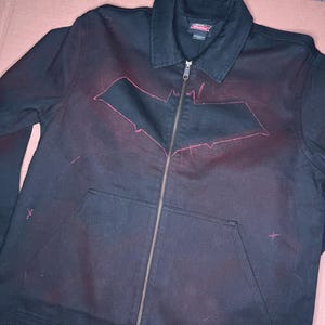 Puede incluir: Chaqueta negra con cremallera y diseño del logotipo de Batman en el centro. La chaqueta tiene cuello, dos bolsillos delanteros y cierre de cremallera. El logotipo de Batman está delineado en rosa rojizo.