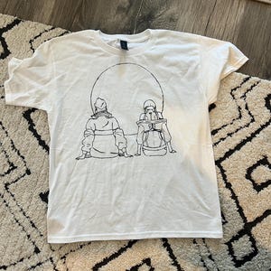 May include: White t-shirt featuring a black line drawing of two figures seated within a circle. The figures are depicted from the back, wearing hooded garments. The shirt is made of a soft, light-colored fabric.