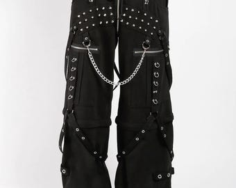 May include: Black cargo pants with multiple pockets, straps, and silver-tone hardware. The pants feature a drawstring waist, a zipper fly, and a chain detail. Studs are placed on the front pockets. The pants are worn with black platform boots.
