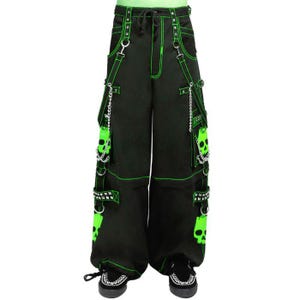 May include: Black wide-leg pants with neon green stitching and accents. The pants feature skull graphics, chains, and studded details. The waist has a drawstring closure and the pants are worn with black sneakers.