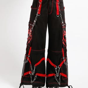 May include: Black cargo pants with red stitching and accents. The pants feature red straps, silver chains, and flame designs. The pants have multiple pockets and zippers, and are worn with black platform shoes.