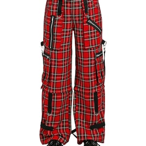 May include: Red plaid cargo pants with black accents, zippers, and straps. The pants have multiple pockets and a drawstring waist. The fabric features a black and white grid pattern over a red background. The pants are worn with black platform shoes.