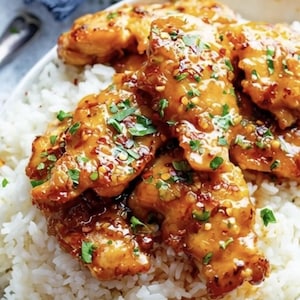 May include: A white bowl filled with white rice and topped with glazed chicken pieces. The chicken is coated in a glossy, orange-brown sauce, sprinkled with red pepper flakes and chopped green herbs. A culinary dish.