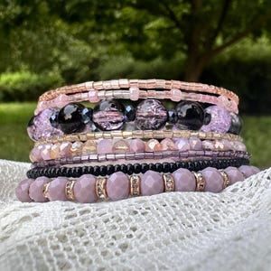 May include: A stack of beaded bracelets in shades of pink, lavender, and black. The bracelets feature various bead sizes and textures, including faceted beads, small seed beads, and larger, round beads. Some bracelets have gold-colored accents.