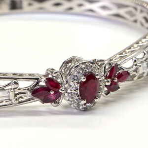 May include: A silver-toned hinged bangle bracelet with an oval ruby center stone surrounded by small white diamonds. The bracelet features additional ruby accents and rectangular white stones.