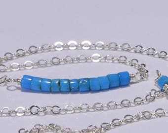Op de afbeelding: Een zilveren ketting met een rij vierkante turquoise blauwe kralen. De ketting is verstelbaar in lengte.