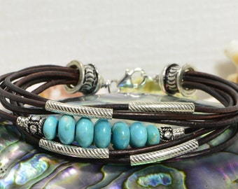May include: A brown leather wrap bracelet with turquoise beads and silver accents. The bracelet is made of multiple strands of leather and has a silver clasp.