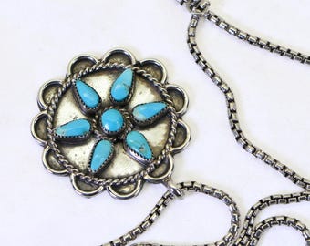 May include: A silver pendant necklace featuring a circular medallion with a scalloped edge and a twisted rope border. The centre is adorned with six teardrop-shaped turquoise stones arranged in a flower pattern around a central round turquoise stone.