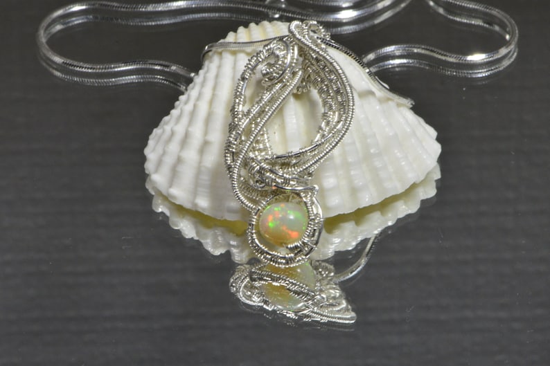May include: A silver wire wrapped pendant with a round, iridescent opal gemstone. The pendant is displayed on a white seashell.