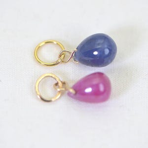 Puede incluir: Dos colgantes de piedras preciosas con aros dorados. Uno tiene forma de lágrima azul oscuro y el otro de lágrima magenta. Los colgantes se muestran sobre una superficie blanca.