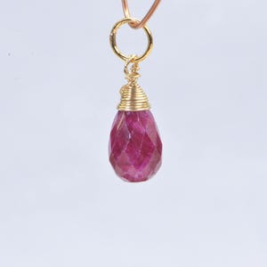 May include: A faceted, teardrop-shaped ruby pendant. The ruby is a deep pinkish-red color and is suspended from a gold-colored ring and wire wrapping. The pendant hangs against a white background.