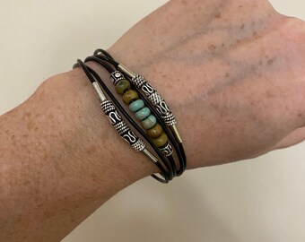 May include: A layered bracelet with black leather cords and silver accents. The bracelet features a row of turquoise and green beads.