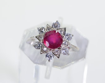 Op de afbeelding: Een zilveren ring met een grote rode edelsteen omringd door kleinere heldere edelstenen in een stervormig ontwerp.