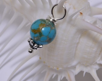 Puede incluir: Un abalorio de cuentas turquesa y dorado con un bucle plateado y detalles decorativos. La cuenta es redonda y tiene un patrón moteado.