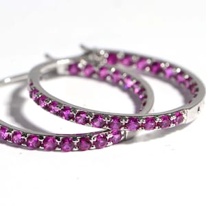May include: A pair of silver hoop earrings, each set with a line of bright, round, pink gemstones. The earrings are displayed against a white background, emphasising the sparkle of the gems and the polished metal.