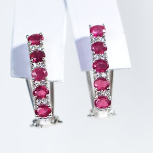 May include: A pair of silver hoop earrings with a row of oval-shaped red gemstones and smaller white gemstones.