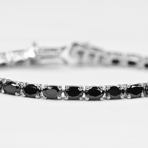 May include: A black diamond tennis bracelet with a silver setting. The bracelet is made of small, round diamonds that are set close together.
