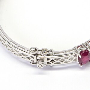 May include: A silver bangle bracelet with a delicate, openwork design. The bracelet is set with a row of oval-shaped ruby gemstones.