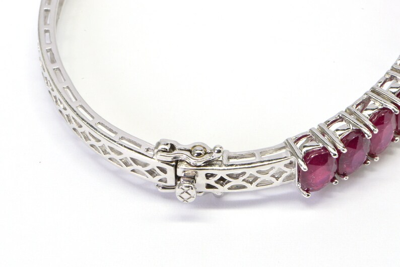 May include: A silver bangle bracelet with a delicate, openwork design. The bracelet is set with a row of oval-shaped ruby gemstones.