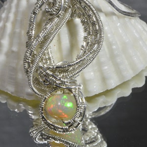 May include: A silver wire wrapped pendant with a round, iridescent opal gemstone. The pendant is displayed on a white seashell.