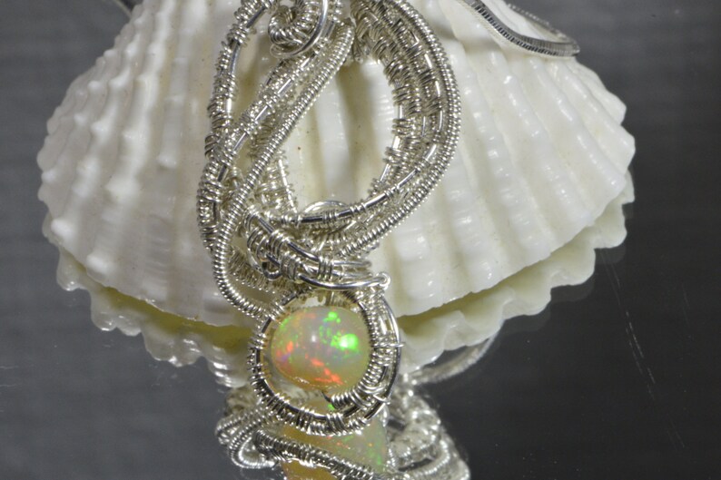 May include: A silver wire wrapped pendant with a round, iridescent opal gemstone. The pendant is displayed on a white seashell.