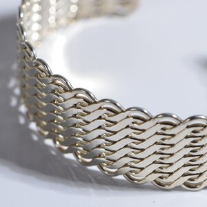 May include: A silver bracelet with a woven design. The bracelet is made of a series of interlocking loops that create a textured surface.