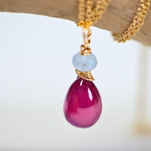 May include: A gold chain necklace with a teardrop-shaped pendant featuring a deep pink gemstone and a smaller light blue gemstone.