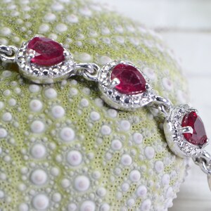 May include: A silver bracelet with red gemstone accents. The bracelet features a series of oval-shaped red gemstones set in silver with a decorative border of smaller white gemstones.