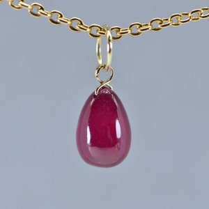 May include: A teardrop-shaped, deep red pendant hangs from a delicate gold chain. The pendant is smooth and polished, reflecting light. The gold chain has small, oval links. The jewelry is set against a soft gray background.
