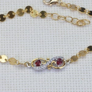May include: Gold-toned bracelet featuring a series of small, flat, circular links. The center of the bracelet showcases two ornate, silver-toned settings, each holding a vibrant red gemstone, surrounded by small, clear stones.