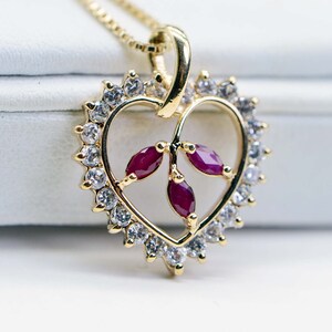 May include: A gold heart-shaped pendant necklace with a delicate vine design and three ruby-red stones. The pendant is surrounded by a halo of clear cubic zirconia stones.