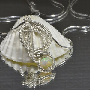 May include: A silver wire wrapped pendant with an iridescent opal gemstone. The pendant is displayed on a white seashell with a silver chain.