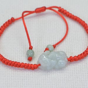 May include: A red string bracelet with a jade green charm in the shape of a Chinese lucky frog. The bracelet has a sliding knot for adjusting the size.