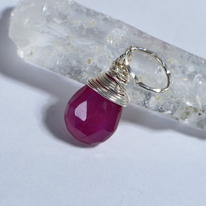 Puede incluir: Un colgante de piedra preciosa rosa envuelto en alambre de plata. El colgante está sobre un cristal de cuarzo transparente.