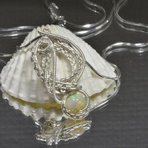 May include: A silver wire wrapped pendant with an iridescent opal gemstone. The pendant is displayed on a white seashell with a silver chain.