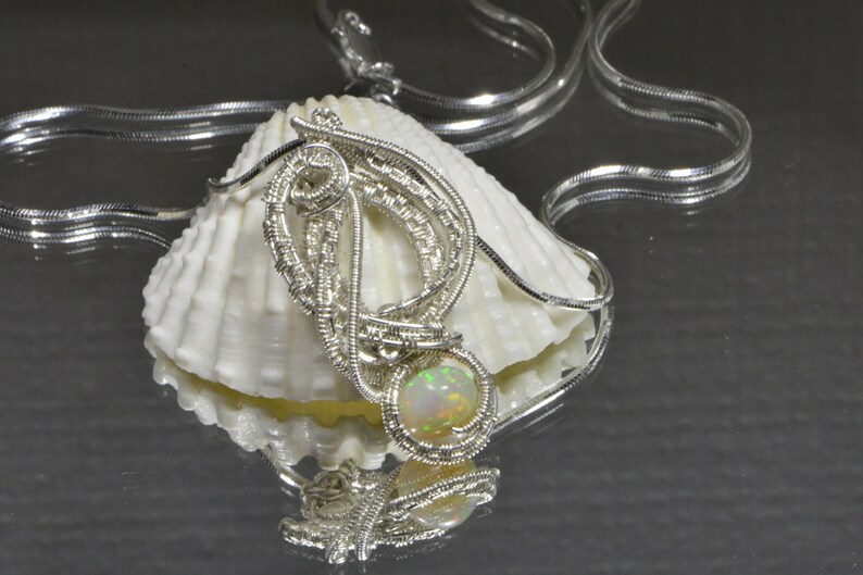 May include: A silver wire wrapped pendant with an iridescent opal gemstone. The pendant is displayed on a white seashell with a silver chain.
